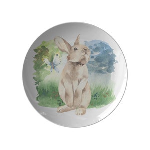 May include: A white ceramic plate with a watercolor illustration of a brown rabbit with a butterfly in a green and blue forest setting.