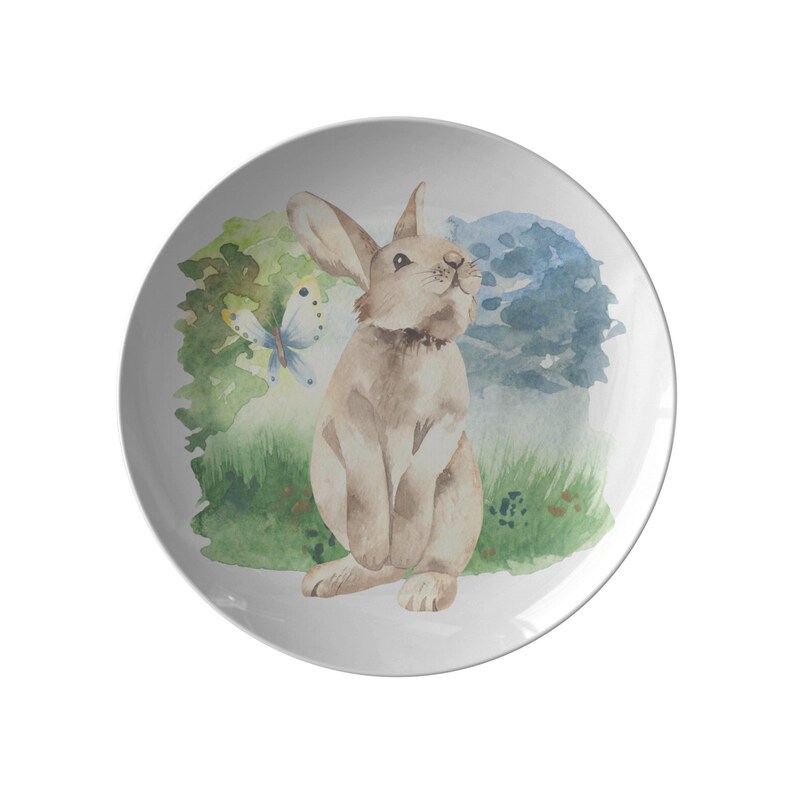 May include: A white ceramic plate with a watercolor illustration of a brown rabbit with a butterfly in a green and blue forest setting.