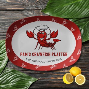 May include: Red oval serving platter with a white border and a cartoon crawfish wearing a chef's hat. The platter has a white outline of crawfish around the border. The text on the platter reads "Pam's Crawfish Platter Let the Good Times Boil."