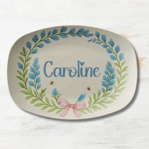 May include: An oval serving platter with a floral design. The platter features a blue floral wreath, a pink bow, and the name "Caroline" in blue script. Two small bees are also included in the design.