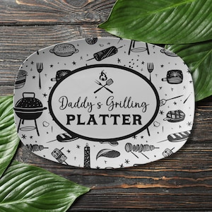 May include: White oval serving platter with a black and white design of grilling tools and food. The center of the platter has a black oval with the text "Daddy's Grilling Platter".