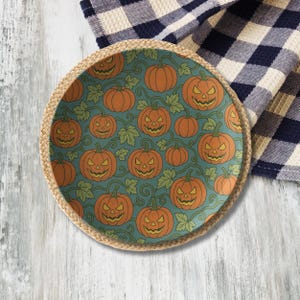 May include: Round decorative plate with a Halloween pumpkin design. The plate features orange jack-o'-lanterns with green vines and leaves on a teal background. The plate has a woven, natural-colored border.