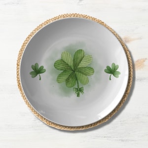 May include: A white ceramic plate with a woven straw placemat. The plate features a green four-leaf clover design with a watercolor effect.