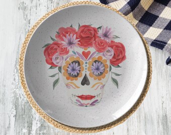 May include: White ceramic plate with a sugar skull design. The skull is decorated with colorful flowers and has a red heart on its forehead. The plate has a speckled white glaze.