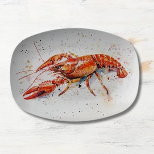 May include: A white oval ceramic plate with a watercolor painting of a red crayfish. The crayfish is painted in a realistic style with details of its claws, antennae, and body.