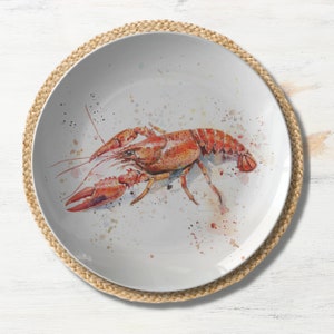 May include: A white ceramic plate with a watercolor illustration of a red crayfish on a white background with colorful watercolor splatters. The plate is on a woven straw placemat.