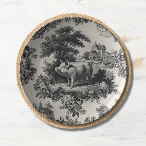 May include: A round decorative plate with a black and white pastoral scene featuring a pig, trees, and a building. The plate has a woven, tan border.