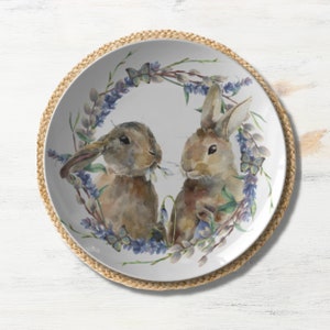 May include: A white ceramic plate featuring two watercolor-style rabbits surrounded by a wreath of lavender and butterflies. The plate rests on a woven placemat, creating a rustic and charming aesthetic. The design is perfect for Easter or spring decor.