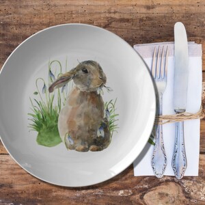 May include: A white ceramic dinner plate with a watercolor illustration of a brown rabbit with blue flowers. The rabbit is surrounded by green grass and blue flowers. The plate is set on a wooden table with a napkin and silverware.