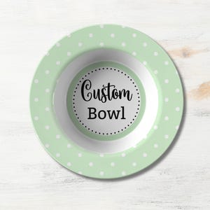 May include: A round bowl with a light mint green rim featuring a white polka dot pattern. The center of the bowl is white and contains the black text Custom Bowl surrounded by a circular border of small black dots on a white background.