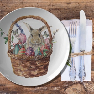 May include: A white ceramic plate with a watercolor illustration of a brown basket filled with Easter eggs, flowers, and a bunny. A fork and knife rest on a white napkin tied with twine.