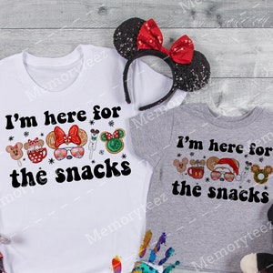 Puede incluir: Dos camisetas blancas con un estampado gráfico negro de personajes de dibujos animados y el texto "I'm here for the snacks". El gráfico incluye un personaje de Mickey Mouse con un gorro de Papá Noel y un personaje de Minnie Mouse con gafas de sol.