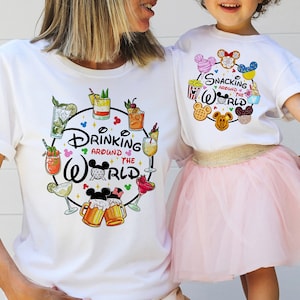 May include: Two white t-shirts with a Disney theme. The adult shirt says "Drinking Around the World" with a circle of Mickey Mouse ears and drinks. The child's shirt says "Snacking Around the World" with a circle of Mickey Mouse ears and snacks.