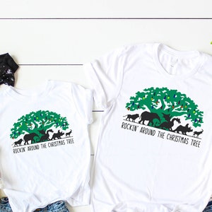 May include: Two white t-shirts with a green tree graphic and the text "Rockin' Around the Christmas Tree." The shirts are paired with denim shorts and white trainers. Accessories include a black bow headband, Minnie Mouse and Mickey Mouse plush toys.