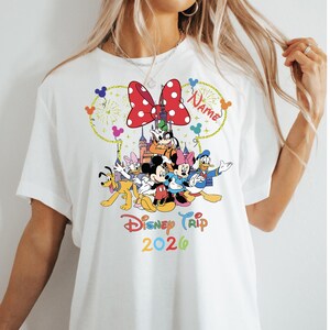 May include: White t-shirt featuring a colorful Disney Trip 2026 design. The graphic includes Mickey and Minnie Mouse, Donald Duck, and other characters, with a red polka-dot bow and fireworks.