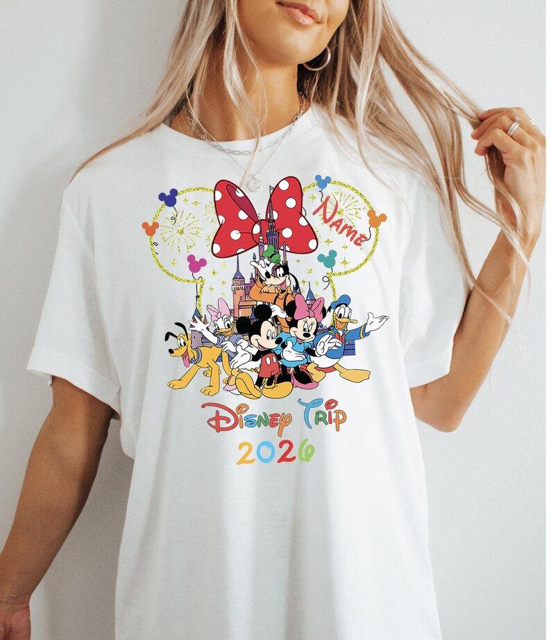 May include: White t-shirt featuring a colorful Disney Trip 2026 design. The graphic includes Mickey and Minnie Mouse, Donald Duck, and other characters, with a red polka-dot bow and fireworks.