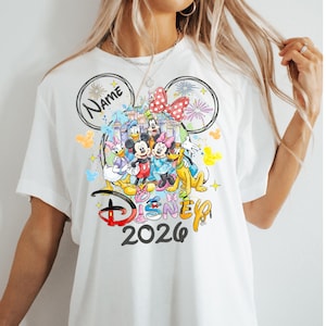 May include: White t-shirt featuring a colorful graphic of Disney characters, including Mickey and Minnie Mouse, Donald Duck, and Goofy. The design includes the text "Disney 2026" and a space for a name, all within a Mickey Mouse ear outline.
