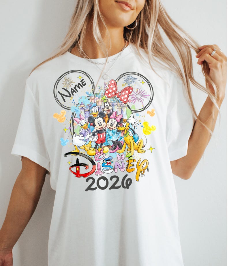 May include: White t-shirt featuring a colorful graphic of Disney characters, including Mickey and Minnie Mouse, Donald Duck, and Goofy. The design includes the text "Disney 2026" and a space for a name, all within a Mickey Mouse ear outline.