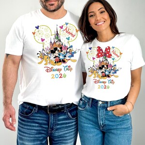May include: White t-shirts featuring a colorful Disney character design. The design includes Mickey Mouse, Minnie Mouse, and other characters, with the text "Disney Trip 2026". The shirts have a round neckline and short sleeves.
