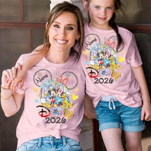 May include: Two light pink t-shirts with a Disney 2026 design. The design features Mickey Mouse, Minnie Mouse, Donald Duck, Goofy, and other characters. The shirts are worn by a woman and a young girl, both smiling.