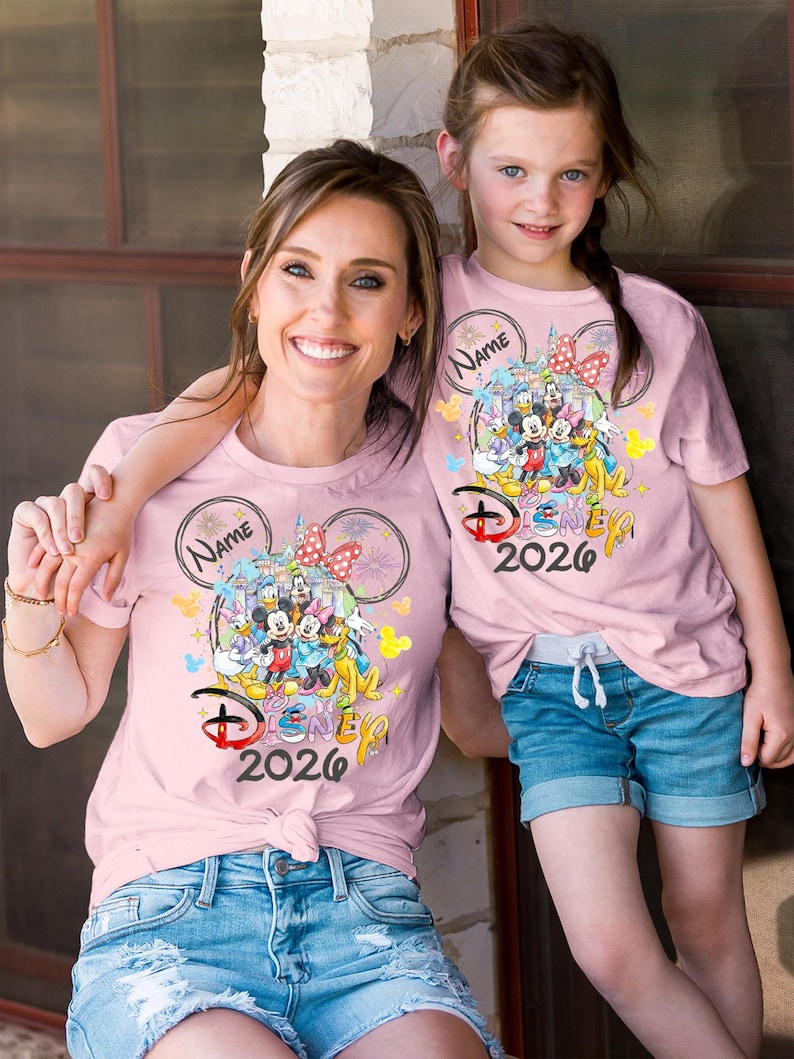 May include: Two light pink t-shirts with a Disney 2026 design. The design features Mickey Mouse, Minnie Mouse, Donald Duck, Goofy, and other characters. The shirts are worn by a woman and a young girl, both smiling.
