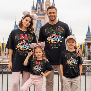 May include: Four people wearing matching black t-shirts with a colorful Disney design and the text "Disney Trip 2026". The shirts feature Mickey Mouse ears, fireworks, and various Disney characters. The family is posing in front of a castle.