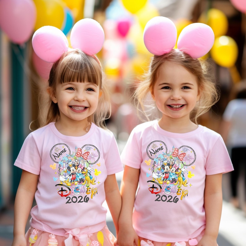 May include: Two young girls wearing matching pink t-shirts with a Disney graphic and the text "Disney 2026". They are also wearing pink balloon headbands. The girls are smiling and holding hands.