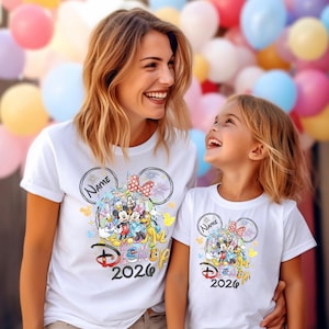 May include: White t-shirts featuring a Disney design with Mickey Mouse, Minnie Mouse, and other characters. The shirts have the text "Disney 2026" and a space for a name. The design includes fireworks and colorful accents.