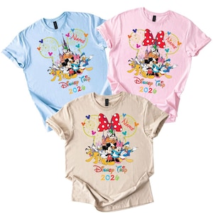 May include: Three short-sleeved t-shirts in light blue, pink, and beige. Each shirt features a colorful graphic of Disney characters, a castle, and the text "Disney Trip 2020". A red polka-dot bow is on the pink and beige shirts.