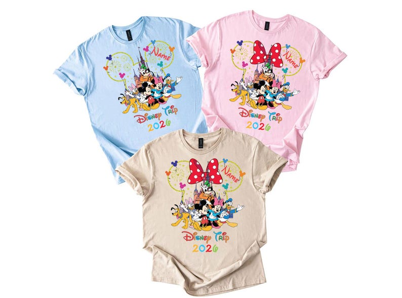 May include: Three short-sleeved t-shirts in light blue, pink, and beige. Each shirt features a colorful graphic of Disney characters, a castle, and the text "Disney Trip 2020". A red polka-dot bow is on the pink and beige shirts.
