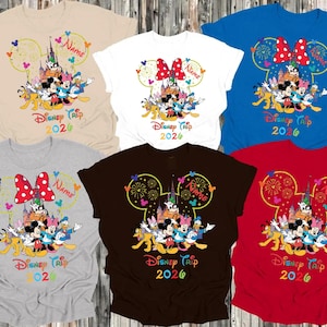 May include: Assorted-color t-shirts featuring a Disney Trip 2026 design. Each shirt displays a Disney character graphic within a Mickey Mouse ear outline, with the text "Disney Trip 2026" below. Colors include beige, white, blue, gray, brown, and red.