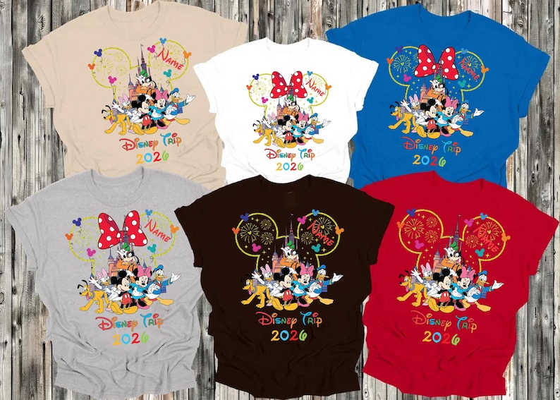 May include: Assorted-color t-shirts featuring a Disney Trip 2026 design. Each shirt displays a Disney character graphic within a Mickey Mouse ear outline, with the text "Disney Trip 2026" below. Colors include beige, white, blue, gray, brown, and red.