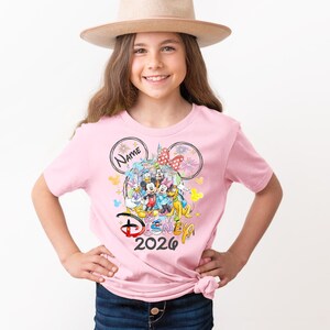 May include: A pink t-shirt featuring a colorful Disney graphic with Mickey and Minnie Mouse, and other characters. The shirt includes the text "Disney 2026" and a space for a name. The girl is wearing a tan hat.