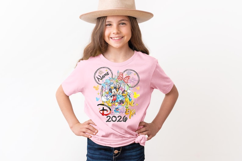 May include: A pink t-shirt featuring a colorful Disney graphic with Mickey and Minnie Mouse, and other characters. The shirt includes the text "Disney 2026" and a space for a name. The girl is wearing a tan hat.