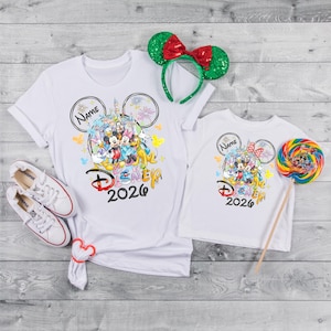 May include: White t-shirts with a colorful Disney graphic, featuring Mickey Mouse and friends, and the text "Disney 2026". A green and red sequined Minnie Mouse ear headband and a colorful lollipop are also present. White sneakers are also in the image.