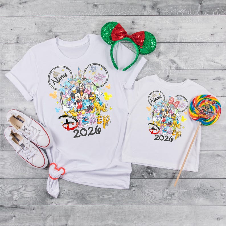 May include: White t-shirts with a colorful Disney graphic, featuring Mickey Mouse and friends, and the text "Disney 2026". A green and red sequined Minnie Mouse ear headband and a colorful lollipop are also present. White sneakers are also in the image.