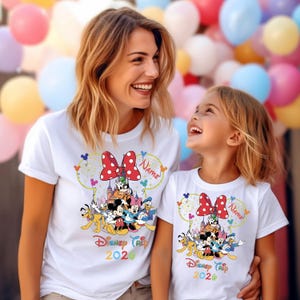 May include: White t-shirts featuring a colorful Disney Trip 2026 design. The design includes a red polka-dot bow, Disney characters, and a castle. The shirts are worn by two people, with a background of colorful balloons.