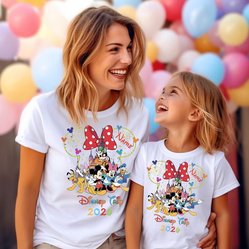 May include: White t-shirts featuring a colorful Disney Trip 2026 design. The design includes a red polka-dot bow, Disney characters, and a castle. The shirts are worn by two people, with a background of colorful balloons.