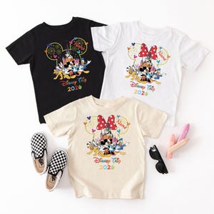 May include: Three short-sleeved t-shirts in black, white, and cream, each featuring a colorful Disney character graphic. The graphic includes Mickey and Minnie Mouse, and the text "Disney Trip 2020". A pair of black and white checkered slip-on shoes is also visible.