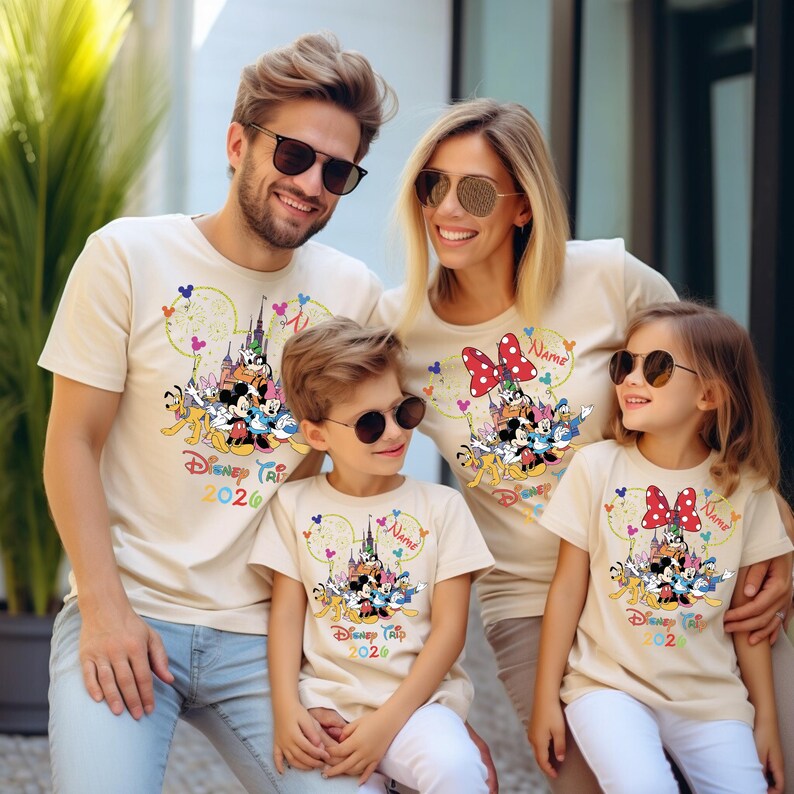 May include: Beige t-shirts with a Disney theme, worn by a family of four. Each shirt features a colorful graphic of Disney characters, a castle, and the text "Disney Trip 2020". The shirts have short sleeves and a crew neck.