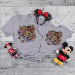 May include: Two grey t-shirts with a floral design featuring the Lion King characters Simba, Nala, Timon, Pumbaa, and Zazu. The shirts are on a wooden background with a pair of Mickey Mouse ears and two plush Mickey Mouse toys.