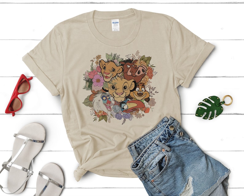 May include: A beige t-shirt featuring a floral design with characters from the Lion King film. The characters include Simba, Nala, Pumbaa, and Timon. The design is in a vintage style with a faded look.