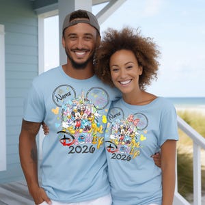 May include: Two people wearing light blue t-shirts with a Disney graphic. The graphic features Disney characters, a castle, and the text "Disney 2026". The shirts have a Mickey Mouse ear design and space for a name.