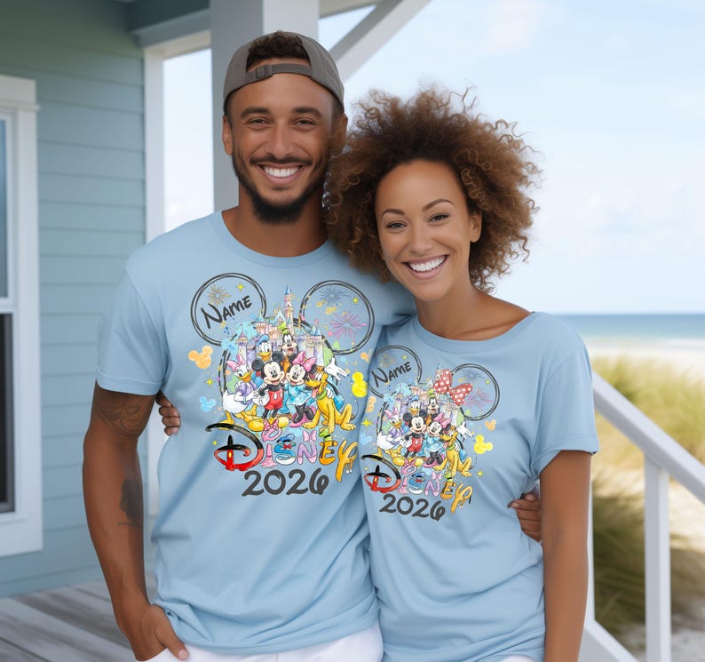 May include: Two people wearing light blue t-shirts with a Disney graphic. The graphic features Disney characters, a castle, and the text "Disney 2026". The shirts have a Mickey Mouse ear design and space for a name.