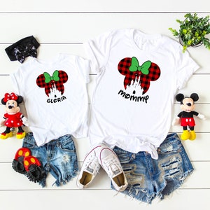 May include: Two white t-shirts with red and green plaid Minnie Mouse ears and the words "mommy" and "Gloria" printed on them. The shirts are on a white wooden surface with a pair of denim shorts, two pairs of shoes, and two stuffed Mickey Mouse toys.