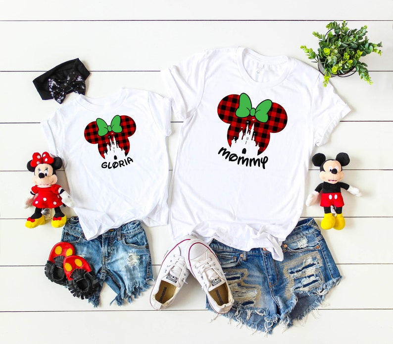 May include: Two white t-shirts with red and green plaid Minnie Mouse ears and the words "mommy" and "Gloria" printed on them. The shirts are on a white wooden surface with a pair of denim shorts, two pairs of shoes, and two stuffed Mickey Mouse toys.