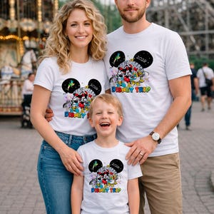 May include: A family of three wearing matching white t-shirts with a colorful Disney graphic. The shirts feature Mickey Mouse, Minnie Mouse, and other Disney characters, with the text "Disney Trip 2020". The family is smiling in front of a carousel.