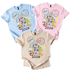 May include: Three short-sleeved t-shirts in light blue, pink, and beige. Each shirt features a colorful graphic of Disney characters, the text "Disney 2026", and space for a name within a Mickey Mouse ear design.