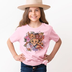 May include: Pink t-shirt with a floral design featuring characters from The Lion King, including Simba, Nala, Pumbaa, and Timon.