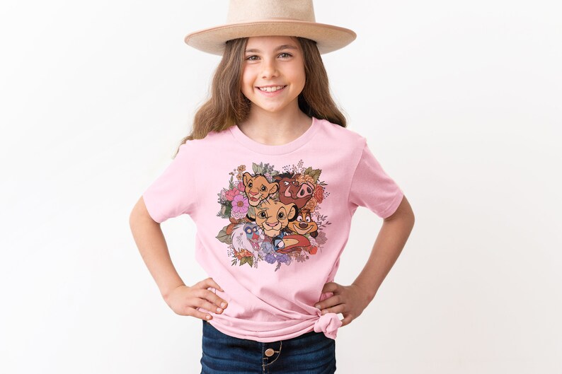 May include: Pink t-shirt with a floral design featuring characters from The Lion King, including Simba, Nala, Pumbaa, and Timon.